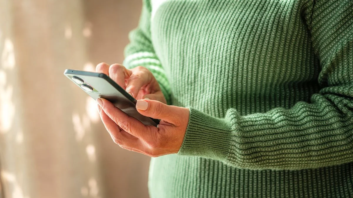 Frau mit grünem Pullover hält ein Smartphone in der einen Hand, während sie es mit dem Zeigefinger der anderen Hand bedient