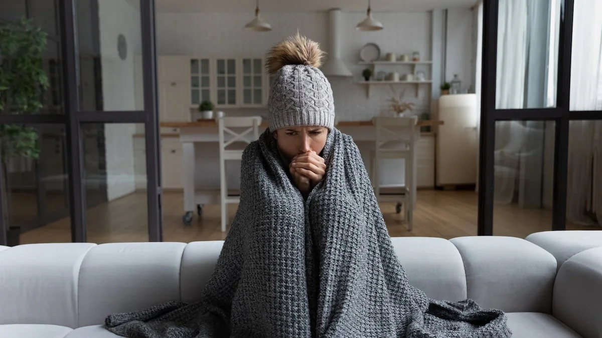 Heizkosten sparen: Junge Frau sitzt frierend mit Mütze und Kuscheldecke in ihrer Wohnung